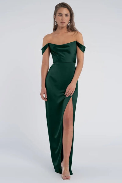 A woman in an elegant off-the-shoulder Sawyer Bridesmaid Dress by Jenny Yoo with a cowl neckline and a thigh-high slit standing against a neutral background.