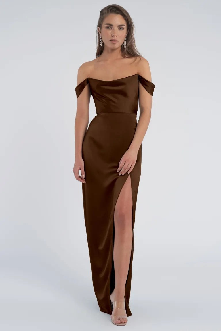 A woman models a luxe Sawyer - Bridesmaid Dress by Jenny Yoo in a satin brown off-shoulder evening gown with a thigh-high slit from Bergamot Bridal.