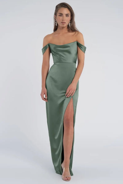 Woman in an elegant green luxe satin Sawyer Bridesmaid Dress by Jenny Yoo with a draped off-the-shoulder neckline and a thigh-high slit from Bergamot Bridal.