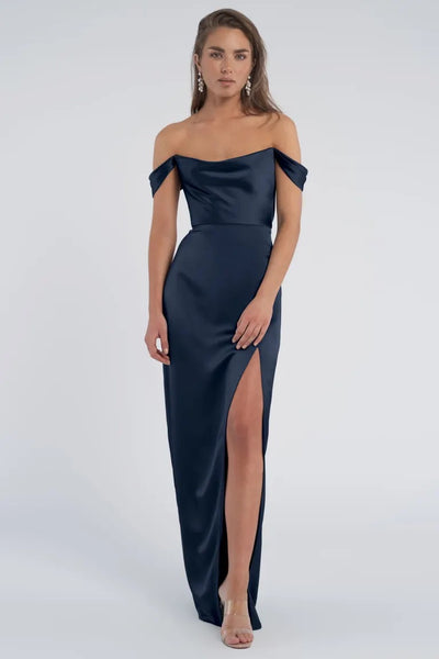 Woman posing in an elegant off-the-shoulder navy blue "Sawyer" bridesmaid dress by Jenny Yoo with a thigh-high slit and cowl neckline from Bergamot Bridal.