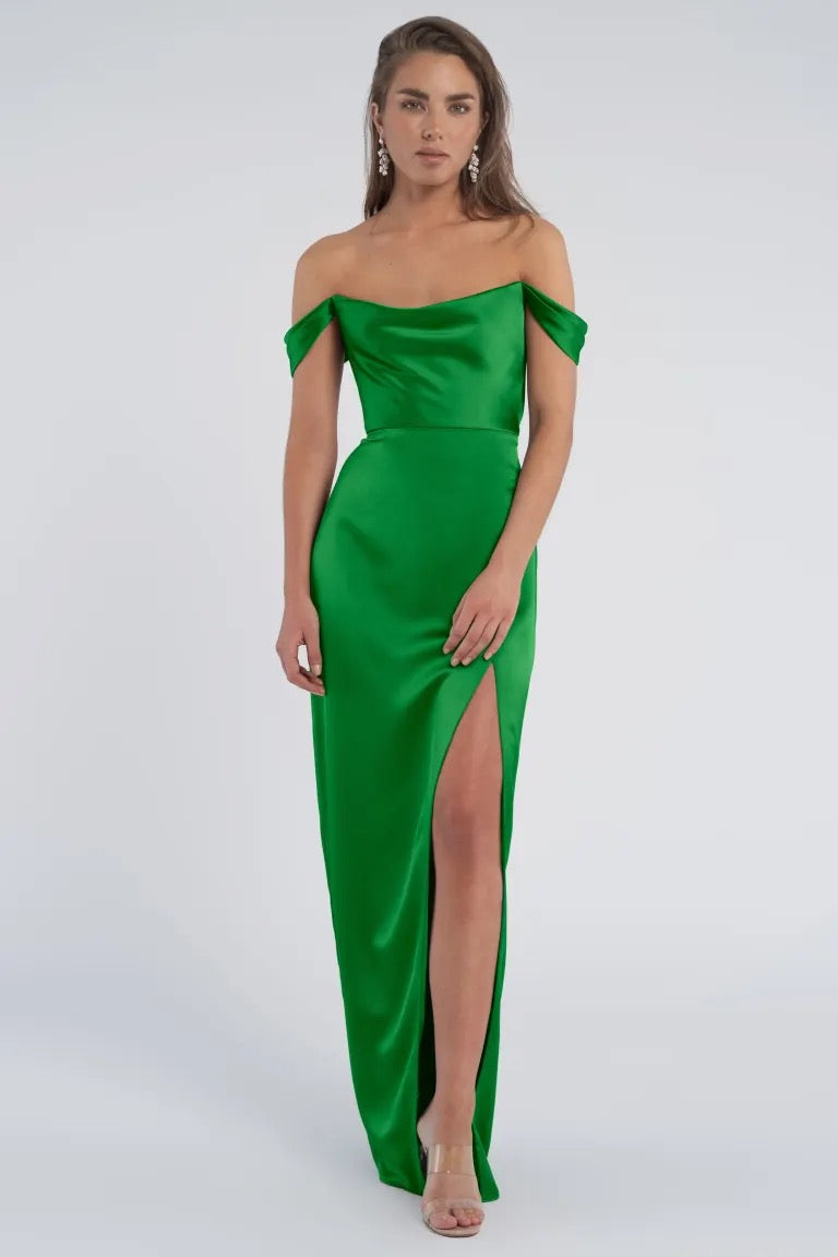 Woman in a Sawyer bridesmaid dress by Jenny Yoo in emerald green satin with an off-the-shoulder design, thigh-high slit, and cowl neckline from Bergamot Bridal.