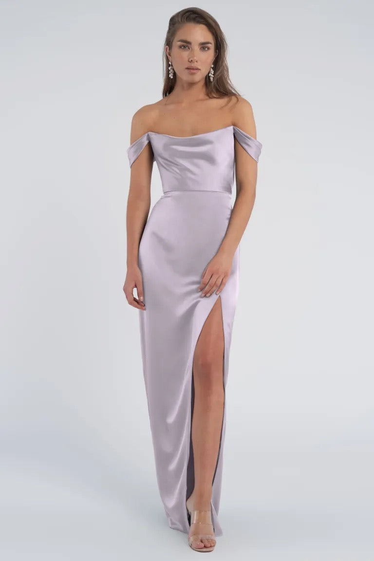 Woman posing in an elegant luxe satin dress with off-the-shoulder sleeves and a thigh-high slit, the Sawyer bridesmaid dress by Jenny Yoo from Bergamot Bridal.