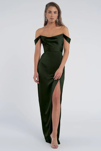 A person stands wearing the Sawyer Bridesmaid Dress by Jenny Yoo from Bergamot Bridal, a luxe satin dark green gown with off-the-shoulder sleeves and a high slit, paired with transparent high heels, against a plain background.