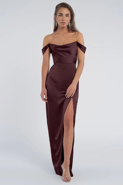 Woman posing in an elegant draped Sawyer - Bridesmaid Dress by Jenny Yoo off-the-shoulder burgundy evening gown with a thigh-high slit from Bergamot Bridal.