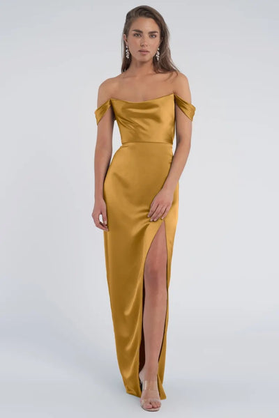 A woman in a luxe satin off-the-shoulder Sawyer - Bridesmaid Dress by Jenny Yoo with a thigh-high slit stands against a plain background.