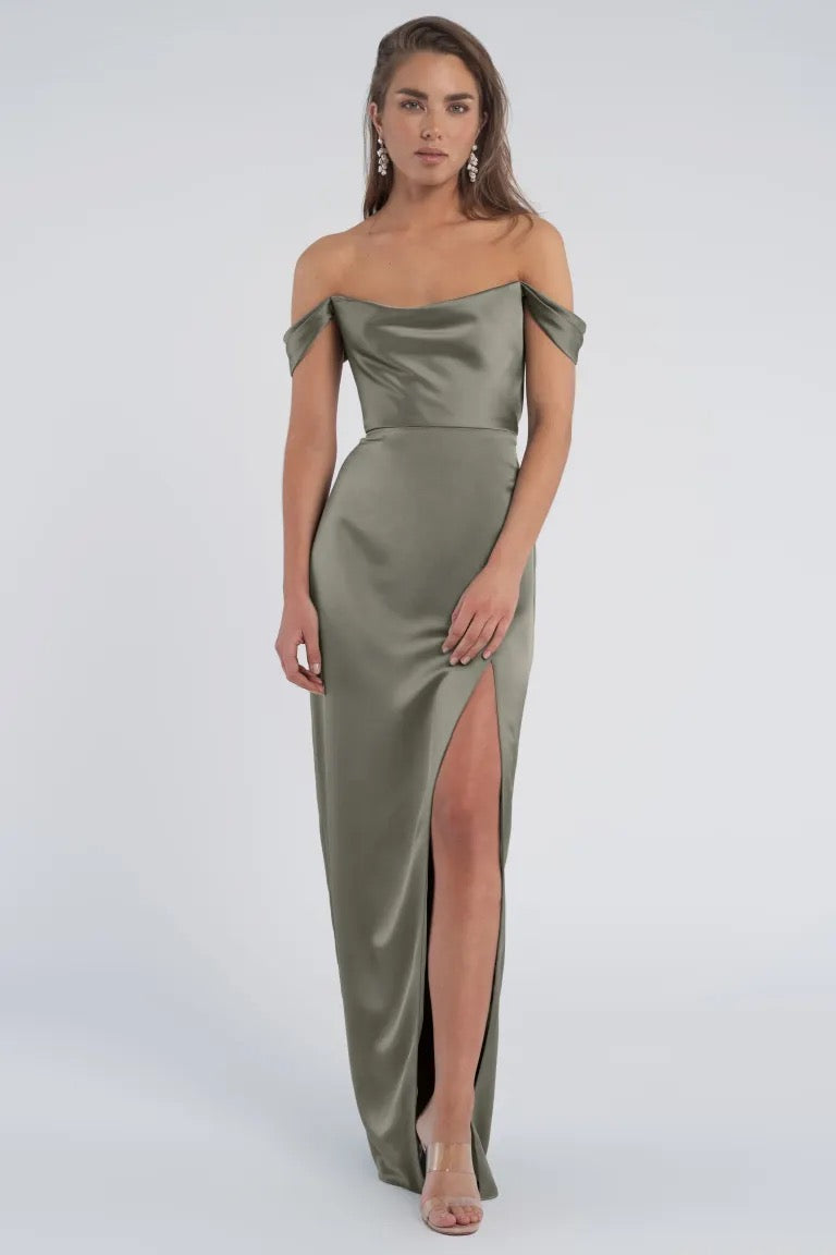 A woman poses in an elegant off-the-shoulder sleeves olive green luxe satin Sawyer Bridesmaid Dress by Jenny Yoo with a thigh-high slit from Bergamot Bridal.