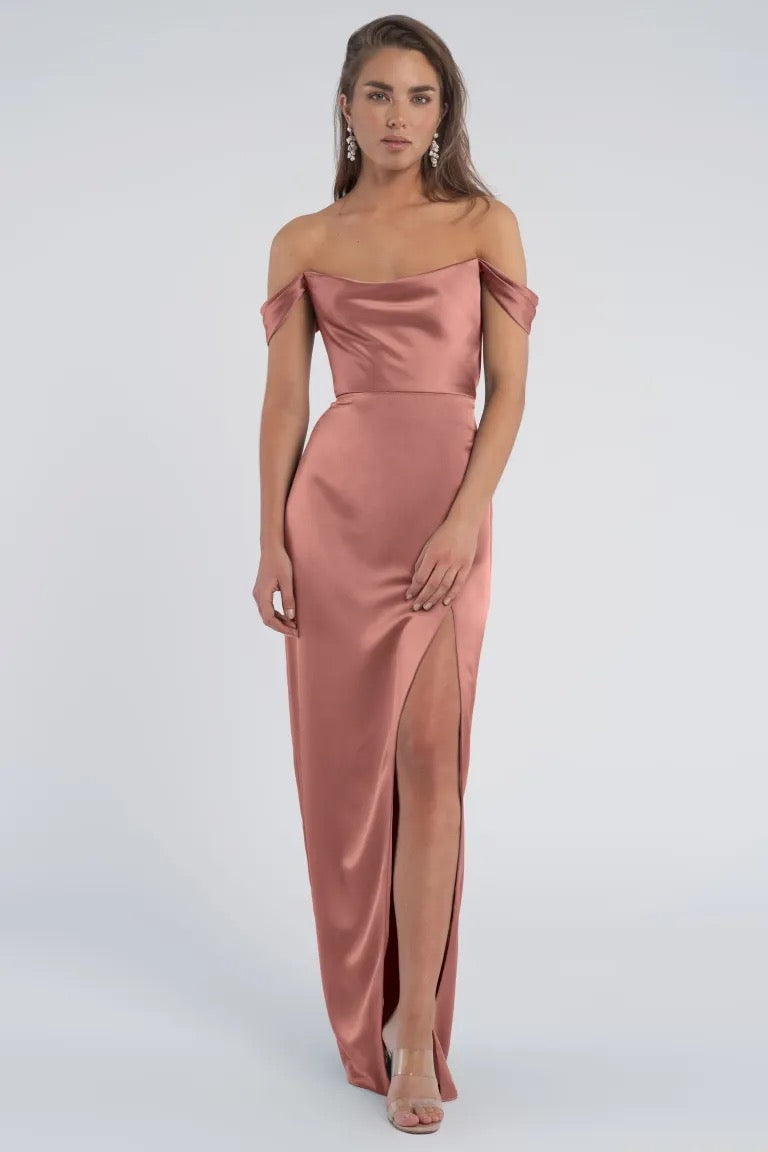 A woman standing in a Sawyer - Bridesmaid Dress by Jenny Yoo from Bergamot Bridal, with an off-the-shoulder, luxe satin design and a thigh-high slit.
