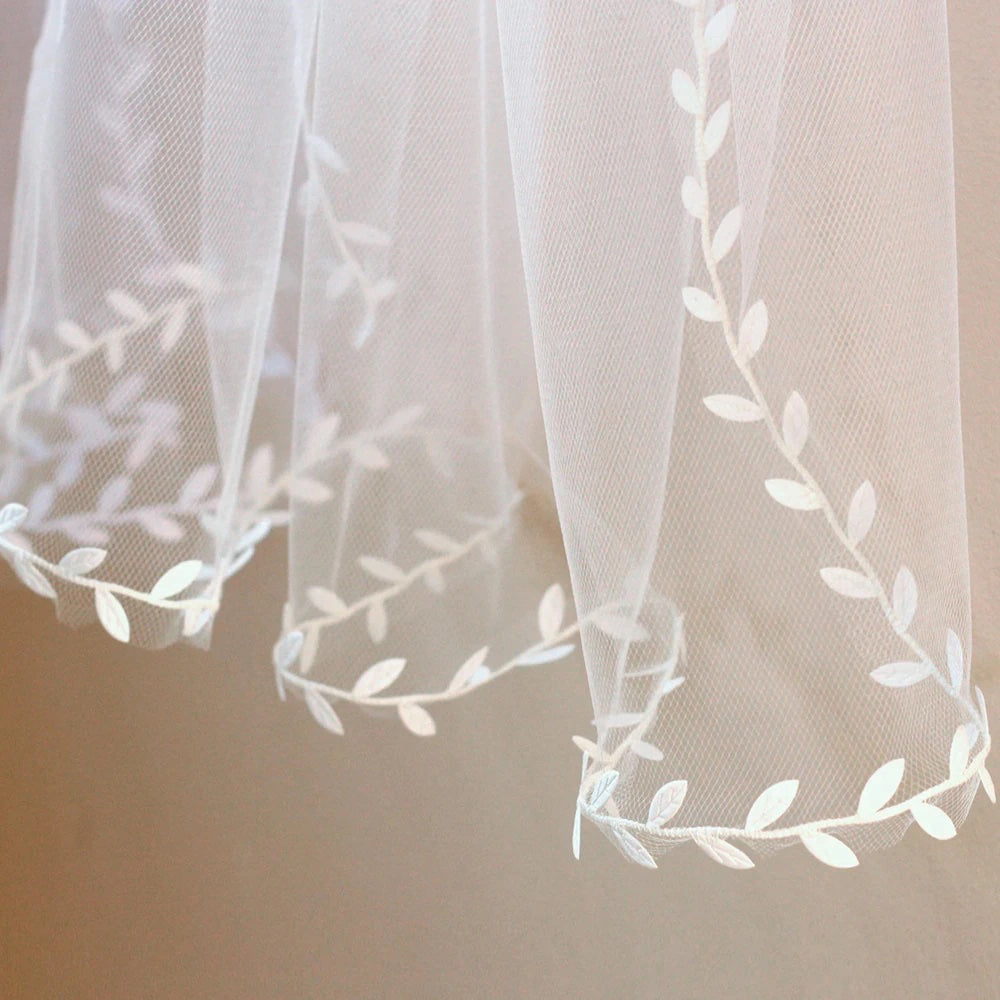 A close-up of the Leaf Lace Edged Bridal Veil by Bergamot Bridal showcases sheer white fabric with a delicate lace leaf design embroidered along its edges, evoking the elegance of a bridal veil.