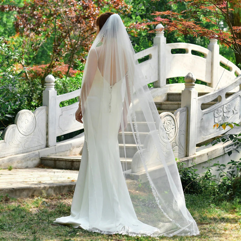 A bride in a white dress, wearing the Bergamot Bridal's "Leaf Lace Edged Bridal Veil" with its delicate lace leaf design and multiple lengths, stands gracefully in front of a decorative stone bridge in the garden.