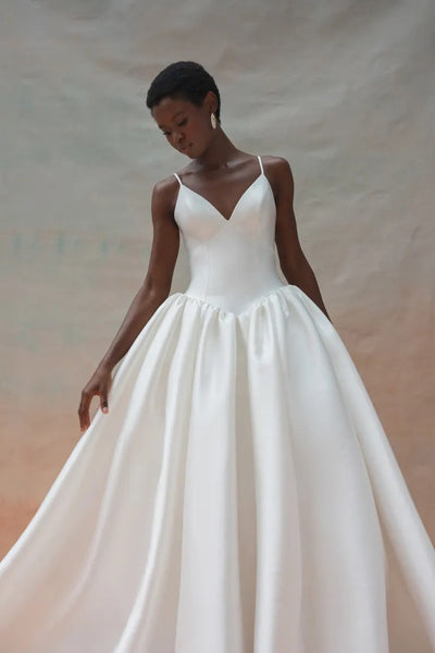 A woman wearing the Shay Wedding Dress by Jenny Yoo from Bergamot Bridal, a floor-length gown made of Mikado fabric with thin straps and a flared skirt, poses against a neutral background.