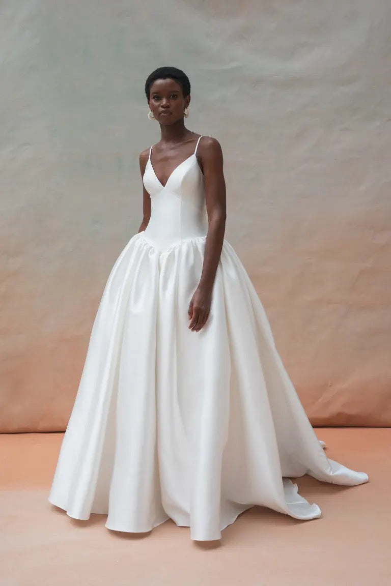 A woman stands against a neutral backdrop, wearing the Shay Wedding Dress by Jenny Yoo from Bergamot Bridal, featuring a sleeveless design with a fitted bodice and a voluminous skirt.