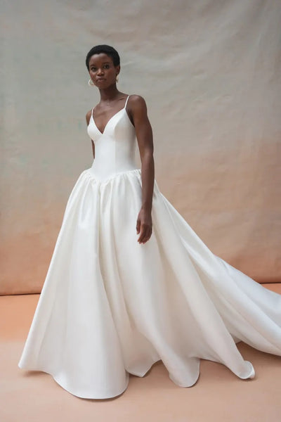 A woman wearing a white, sleeveless Shay Wedding Dress by Jenny Yoo from Bergamot Bridal, featuring a V-neckline and a long, flowing skirt crafted from luxurious Mikado fabric, stands against a neutral backdrop.