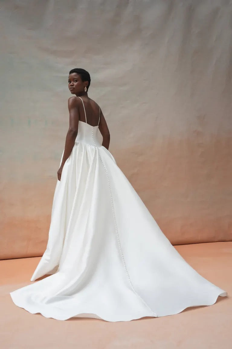 A person with short hair is elegantly dressed in the Shay Wedding Dress by Jenny Yoo from Bergamot Bridal, crafted from luxurious Mikado fabric. The gown features a long train and a Basque waist, and the individual is standing sideways against a plain gradient background of beige and peach tones.