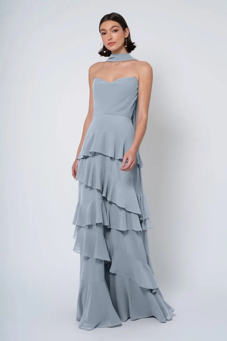 A woman posing in a strapless cowl neckline, tiered grey Jenny Yoo Simona chiffon bridesmaid dress against a white background from Bergamot Bridal.