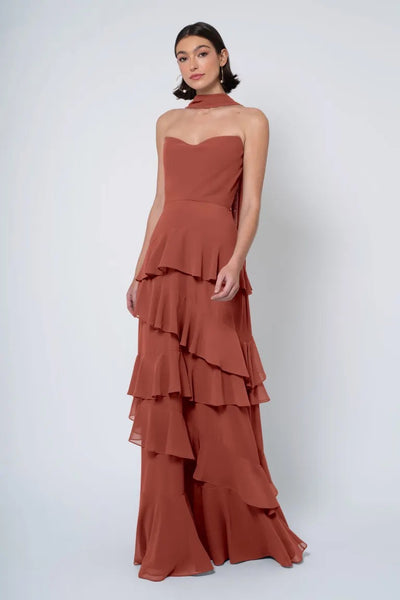 A woman in an elegant, strapless cowl neckline terracotta gown with a ruffled skirt posing against a light background, wearing the Simona bridesmaid dress by Jenny Yoo from Bergamot Bridal.