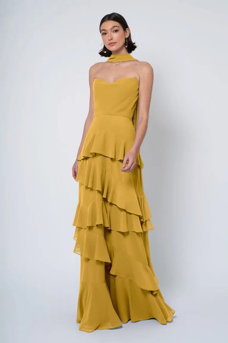 A woman poses in an elegant tiered mustard yellow Simona - Jenny Yoo Bridesmaid Dress with a strapless cowl neckline from Bergamot Bridal.