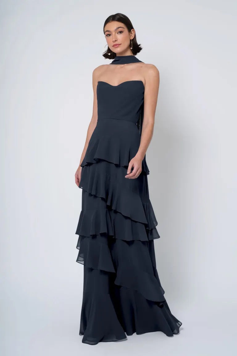 A woman in a Simona - Jenny Yoo Bridesmaid Dress from Bergamot Bridal, with a strapless cowl neckline, black tiered gown posing against a white background.