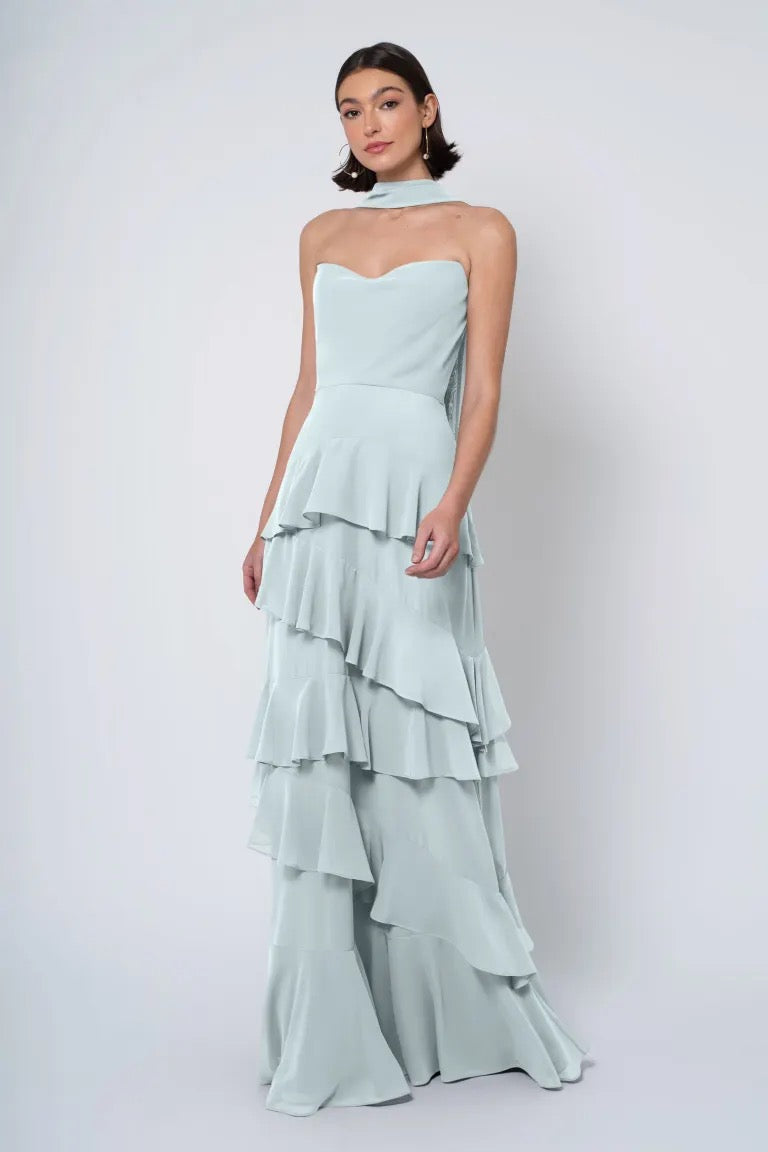 A woman posing in a Simona - Jenny Yoo Bridesmaid Dress by Bergamot Bridal, a light blue strapless cowl neckline gown with a ruffled skirt against a white background.