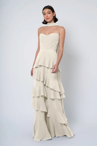 A woman stands posing in an elegant, strapless Simona - Jenny Yoo bridesmaid dress with a tiered ruffled skirt against a light background from Bergamot Bridal.