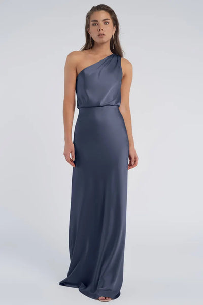 A woman wearing a one-shoulder neckline Jenny Yoo Bridesmaid dress in luxe satin fabric stands against a neutral background from Bergamot Bridal.