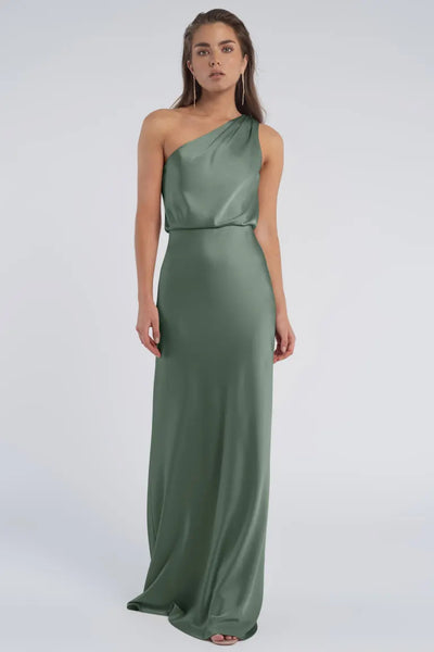 A woman in an elegant green Jenny Yoo Bridesmaid dress with a one-shoulder neckline stands against a neutral background.