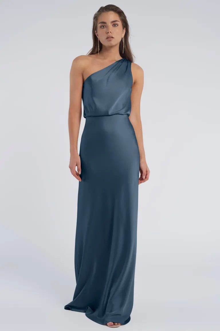 A woman in an elegant blue one-shoulder Sterling - Jenny Yoo Bridesmaid dress with a blouson waist stands against a neutral background from Bergamot Bridal.