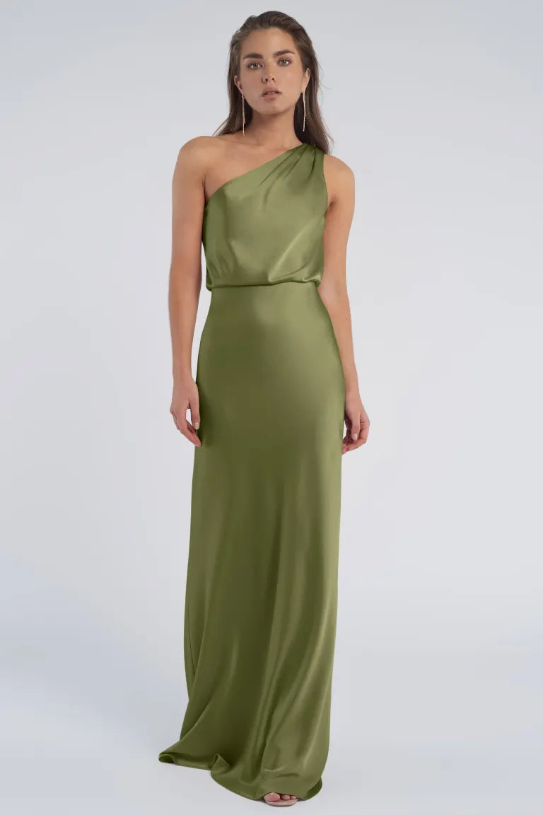 A person is shown wearing the "Sterling" Jenny Yoo Bridesmaid gown by Bergamot Bridal, a sleek, olive green, one-shoulder, floor-length dress made of luxe satin fabric, standing against a plain light-colored background.