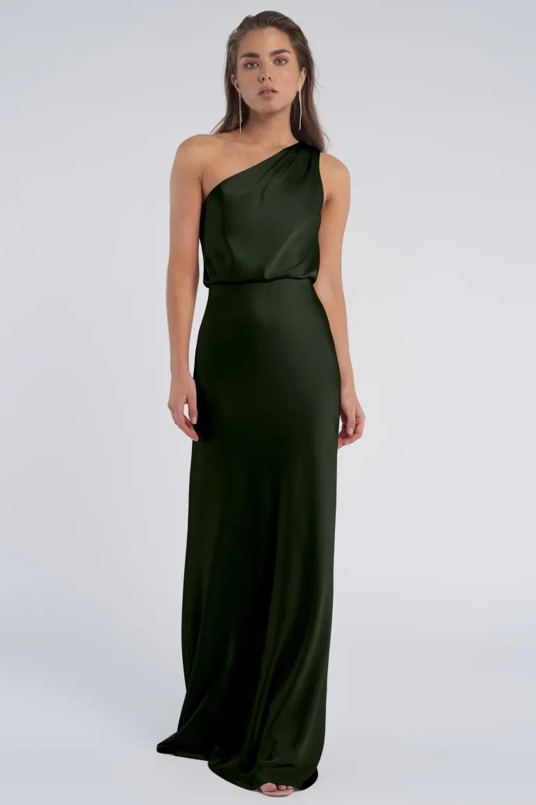 A person stands wearing the Sterling dress from Bergamot Bridal, made of dark green luxe satin fabric, featuring a one-shoulder neckline and floor-length elegance.