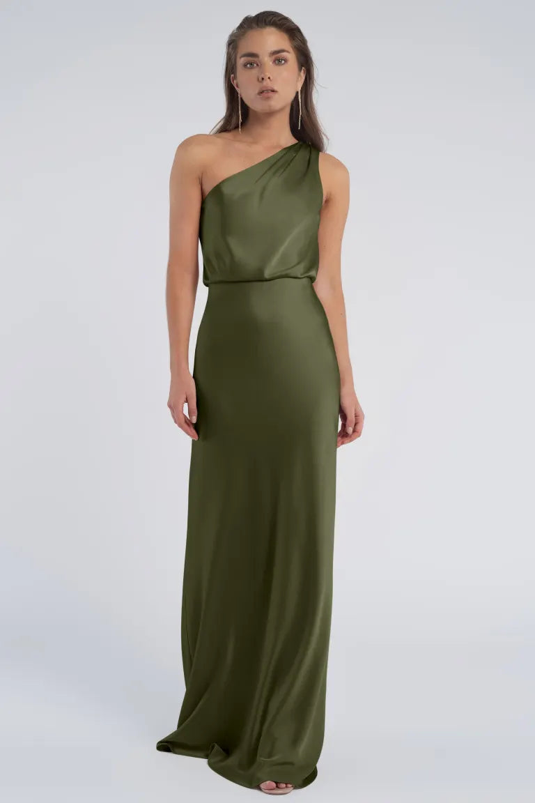 A woman in an elegant green Sterling - Jenny Yoo Bridesmaid dress with a one-shoulder neckline standing against a neutral background. (Brand Name: Bergamot Bridal)