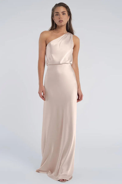 A woman standing against a neutral background, wearing an elegant, one-shoulder neckline, long beige luxe satin Sterling dress by Jenny Yoo Bridesmaid.