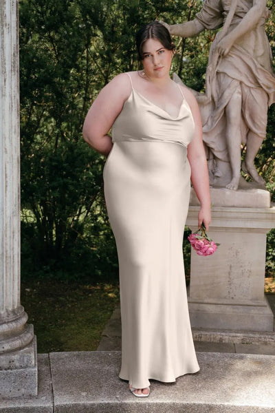 A woman in a Sylvie - Bridesmaid Dress by Jenny Yoo from Bergamot Bridal holding a bouquet stands next to a statue in a garden setting, exuding Old Hollywood vibes.