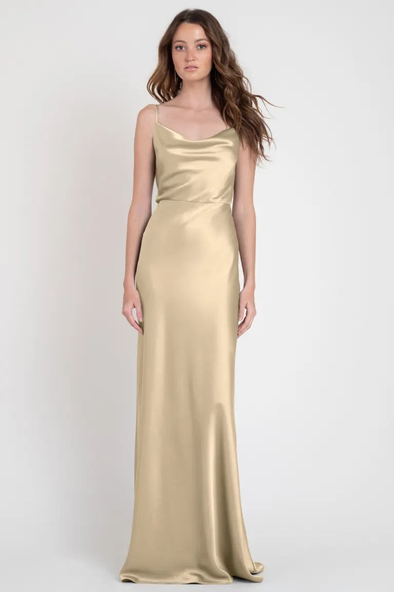 A woman standing in a studio wearing an elegant gold satin Sylvie - Bridesmaid Dress by Jenny Yoo from Bergamot Bridal.