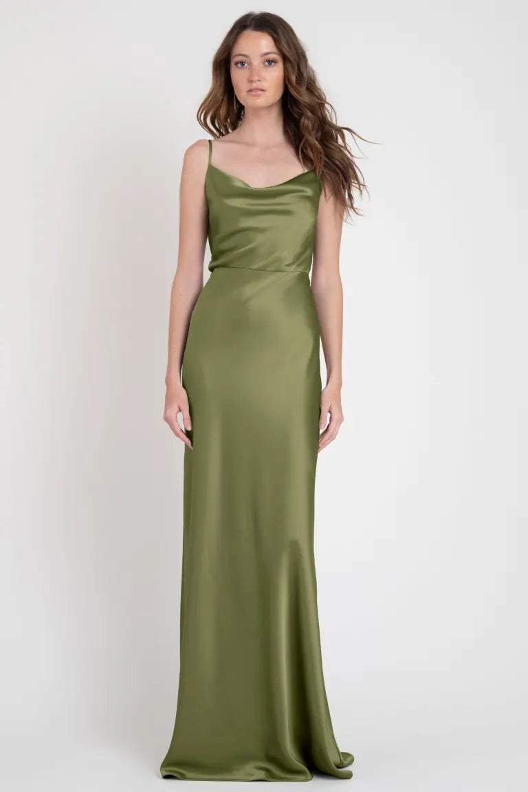 A woman stands in a studio wearing the Sylvie Bridesmaid Dress by Jenny Yoo for Bergamot Bridal. The long, sleeveless, olive green satin slip dress enhances her elegant hairstyle of loose waves, exuding an Old Hollywood allure.