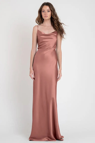A woman standing in a studio wearing an elegant Sylvie blush satin slip dress by Jenny Yoo, exuding Old Hollywood vibes from Bergamot Bridal.