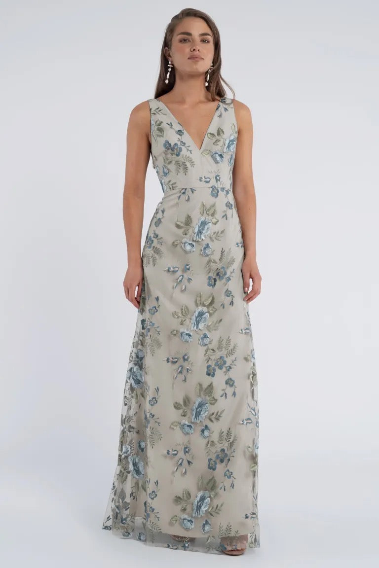 A woman is standing and wearing the Tatum Bridesmaid Dress by Jenny Yoo from Bergamot Bridal, a long, sleeveless V-neck dress adorned with blue and green floral patterns against a plain background. The dress features an elegant slim A-line skirt. She has long hair and is looking straight ahead.