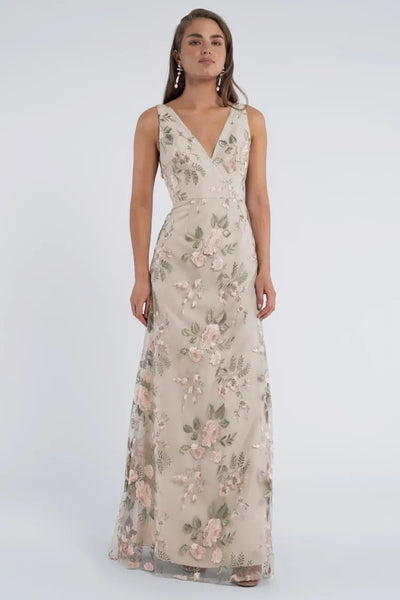 A woman wearing the Tatum Bridesmaid Dress by Jenny Yoo, a sleeveless, floor-length gown made of Enchanted Floral embroidered tulle fabric from Bergamot Bridal, stands against a plain background.