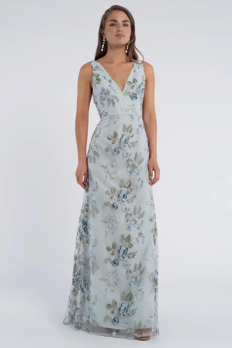 A person stands wearing the Tatum Bridesmaid Dress by Jenny Yoo, a long, sleeveless gown with a V-neckline featuring a light blue floral print from Bergamot Bridal. They have long hair and wear dangling earrings.