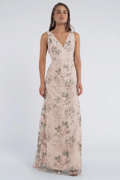 A woman wears a sleeveless Tatum Bridesmaid Dress by Jenny Yoo, featuring Enchanted Floral embroidered tulle fabric on a light background. She stands against a plain white backdrop. The dress, by Bergamot Bridal, showcases intricate floral details perfect for a bridesmaid look.