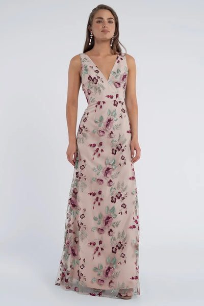 A woman wearing the Tatum Bridesmaid Dress by Jenny Yoo from Bergamot Bridal, featuring a floor-length, sleeveless V-neck design with a slim A-line skirt and floral pattern, stands elegantly against a plain backdrop.