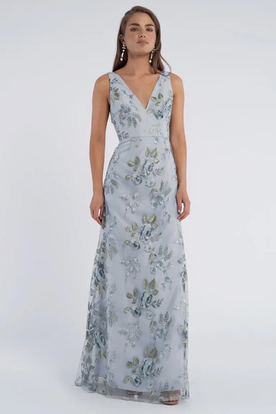 A person stands wearing the captivating, floor-length Tatum Bridesmaid Dress by Jenny Yoo from Bergamot Bridal, featuring a sleeveless design in light blue with a floral pattern and a V-neckline. The intricate details of the Enchanted Floral embroidered tulle fabric make it perfect for any elegant occasion.