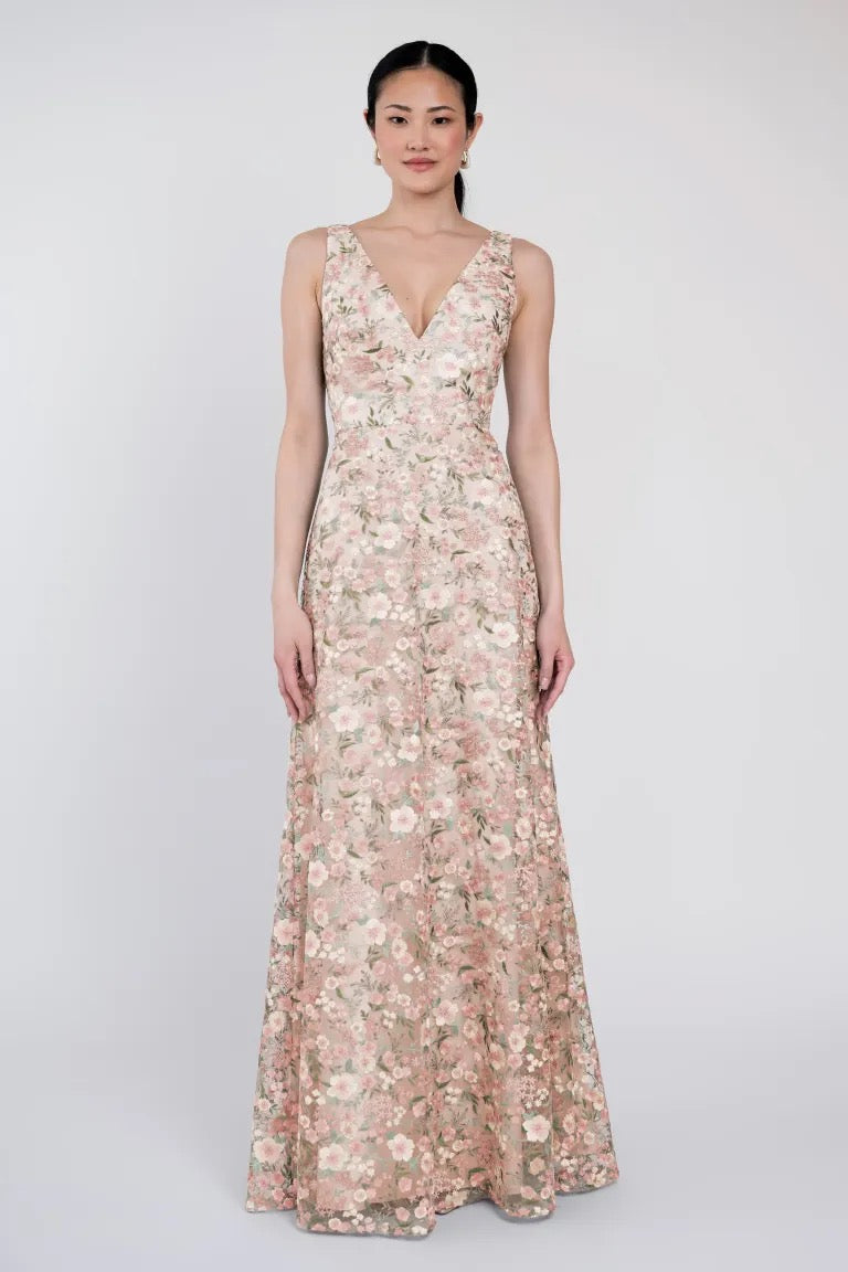 A woman stands wearing an enchanting sleeveless V-neck, floral-patterned, floor-length dress reminiscent of the Tatum Bridesmaid Dress by Jenny Yoo from Bergamot Bridal, against a neutral background.