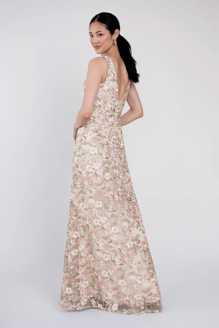 A woman stands against a plain background wearing the Tatum Bridesmaid Dress by Jenny Yoo from Bergamot Bridal, featuring a deep V-back. She glances over her shoulder towards the camera, her hair tied in a low ponytail.