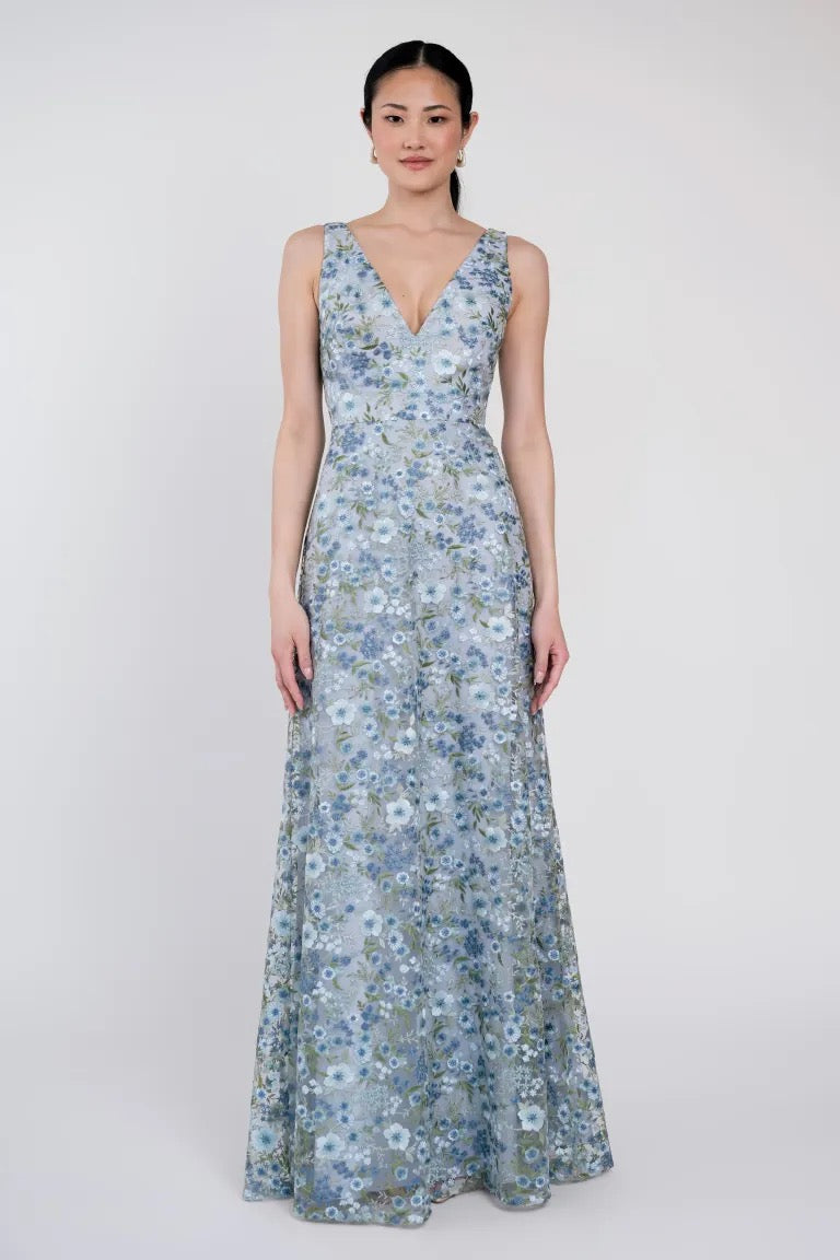 A woman stands wearing the Tatum Bridesmaid Dress by Jenny Yoo, a floor-length, sleeveless gown with a floral pattern and a slim A-line skirt that adds elegance to the V-neck design from Bergamot Bridal.