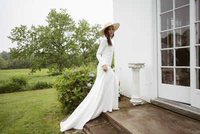 A woman in the Taylor by Jenny Yoo gown from Bergamot Bridal, featuring elegant long sleeves, stands outdoors near a white building with greenery, flowers, and a grassy field in the background.