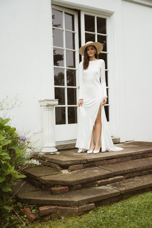 A woman in the Taylor by Jenny Yoo gown from Bergamot Bridal, with elegant long sleeves and a wide-brimmed hat, stands on stone steps before glass doors beside a broken white column.
