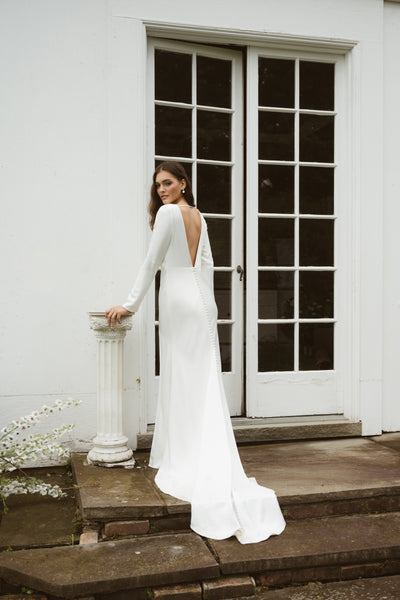A woman in the Taylor by Jenny Yoo gown from Bergamot Bridal, featuring elegant long sleeves and an open back, stands on stone steps before glass-paneled doors, resting her hand on a white column.