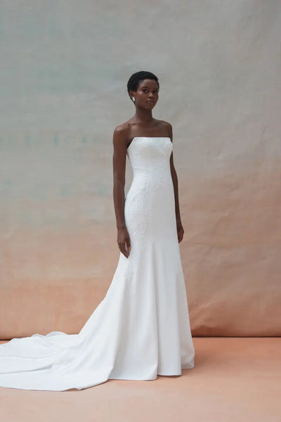 A woman stands wearing the Tricia Wedding Dress by Jenny Yoo from Bergamot Bridal, featuring a strapless white design with a long train and fit and flare silhouette, in front of a light-colored background.