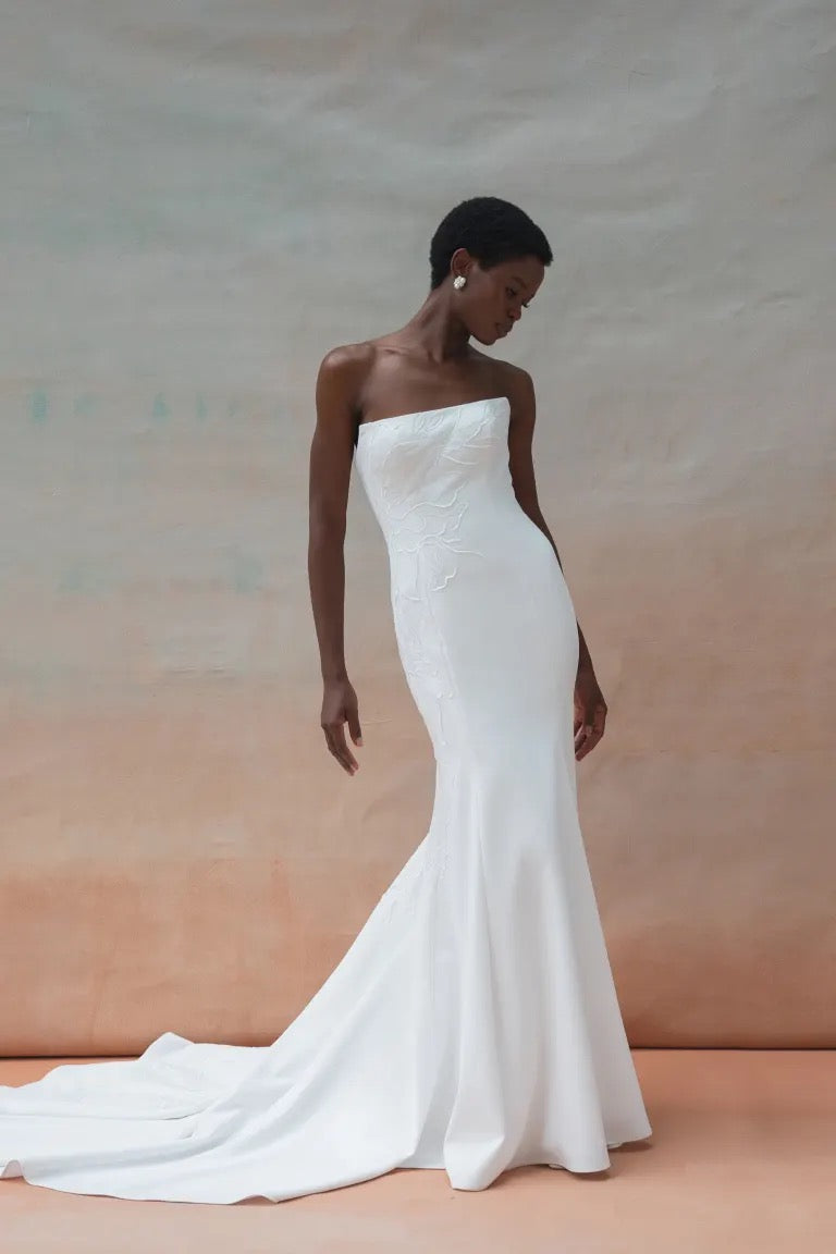 A woman in a strapless white Tricia Wedding Dress by Jenny Yoo from Bergamot Bridal poses against a neutral background. The dress, with its fit and flare silhouette, features a fitted bodice adorned with floral organza applique and a flowing train.