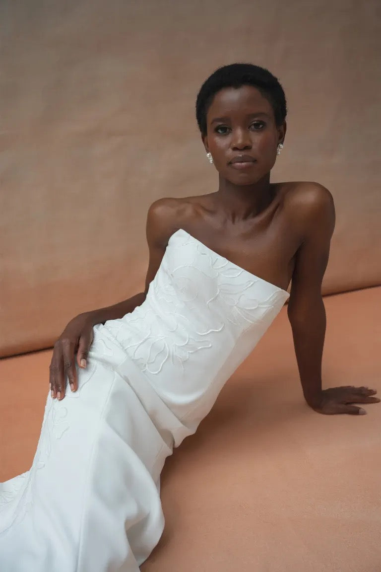 A woman wearing the Tricia Wedding Dress by Jenny Yoo from Bergamot Bridal, adorned with floral organza applique, is seated on the floor with one hand resting on it. The fit and flare silhouette complements the neutral background beautifully.