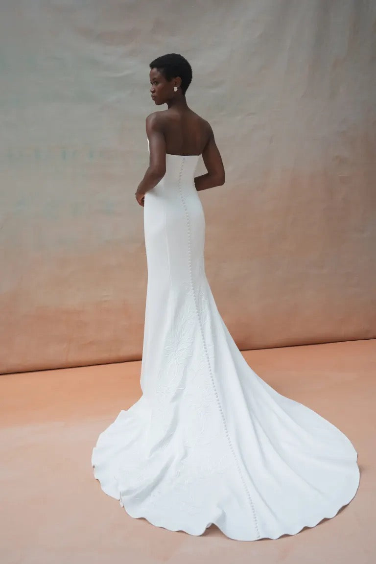 A woman in a strapless white Tricia Wedding Dress by Jenny Yoo from Bergamot Bridal poses against a textured background. The gown features exquisite floral organza applique and a fit and flare silhouette, and she is turned slightly to her side, showcasing the back and long train.